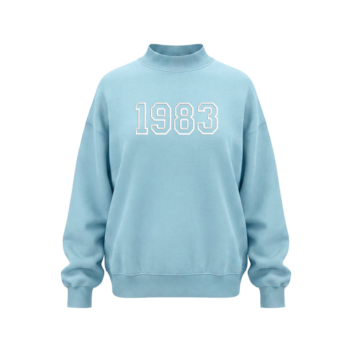 A light blue mock neck sweatshirt with '1983' embroidered in white varsity-style block letters across the chest, displayed on a clean white background.