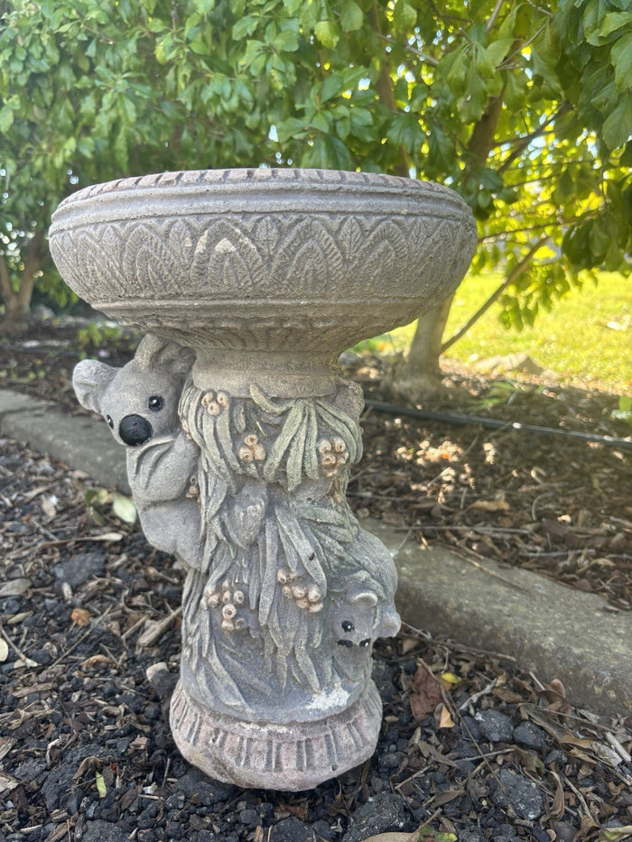 concrete bird bath featuring a koala and possum design in a natural garden setting