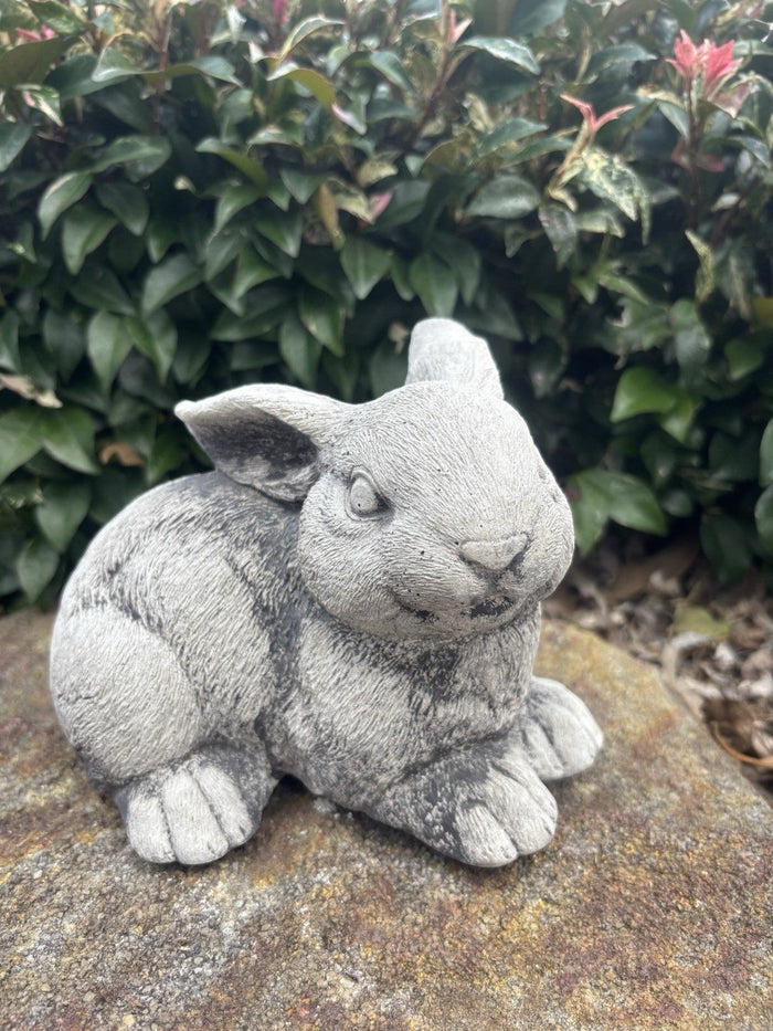 handcrafted grey concrete rabbit garden ornament sitting on a stone in a garden setting