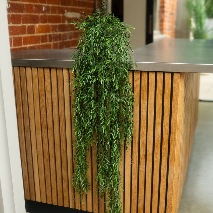 A green trailing houseplant with dense leafy foliage draped over a modern wooden slat counter in an indoor setting with a brick wall and shelving in the background.