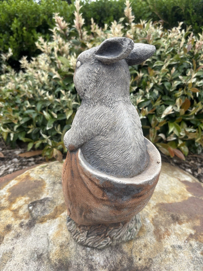 22nice4me Garden Statues hand-painted concrete rabbit sitting in a potato sack, back view in a garden setting.