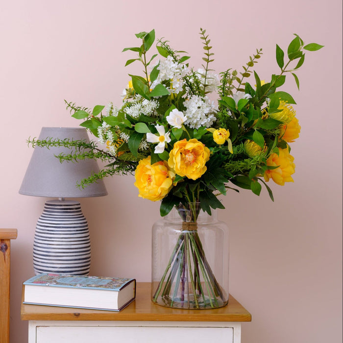Floral arrangement in a vase on a nightstand next to a bed with a lamp and book.