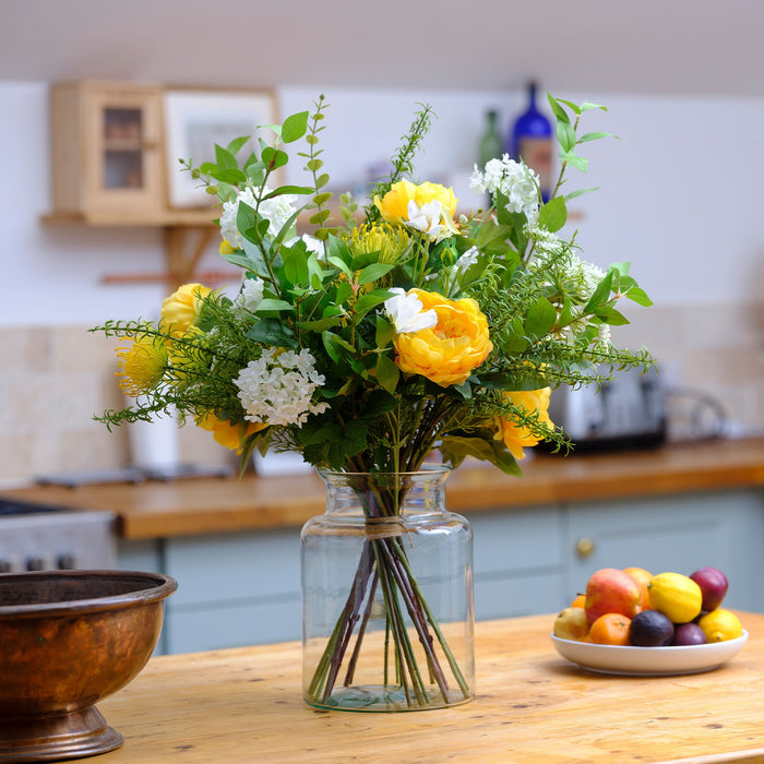 Bouquet of yellow, white, and green flowers in a glass vase on a wooden table with a kitchen background.