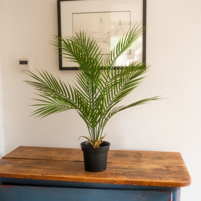 A potted artificial green palm plant in a black pot, displayed on a rustic wooden table against a white wall with a framed print.