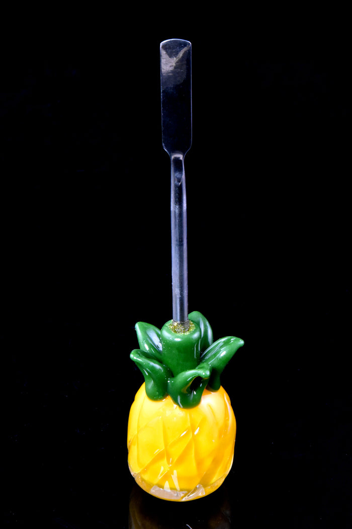 Pineapple-shaped glass dab tool with stainless scoop tip standing upright on glossy black background, showing faceted yellow body and green crown.