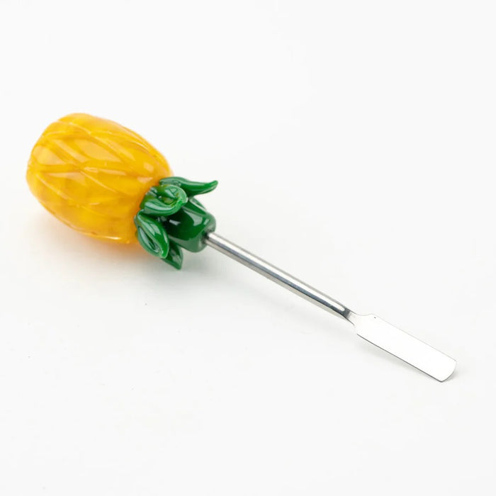 Angled side shot of pineapple dab tool lying down, focusing on detailed rind texture, green crown, and full-length metal spatula tip on white surface.